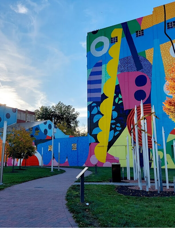 A colorful abstract mural covers the side of a building next to a park path lined with metal poles, signs, green grass, and small trees under a blue sky.