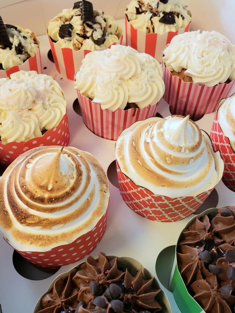 A close-up of assorted cupcakes in colorful paper wrappers, topped with swirls of frosting, toasted meringue, chocolate chips, and cookie pieces, arranged in a box.