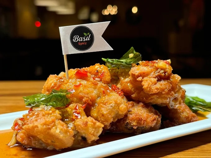 A plate of crispy fried chicken pieces coated in a glossy, spicy sauce, garnished with fresh basil leaves and topped with a small flag labeled 