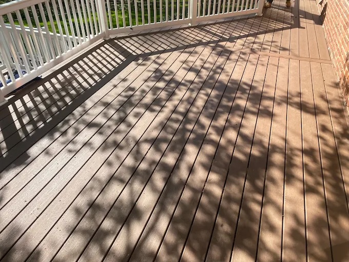 Sunlight casts tree branch shadows across a clean, brown wooden deck with white railings. The deck is attached to a brick building and overlooks a green lawn.
