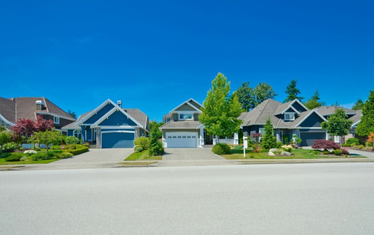 A row of modern suburban houses with well-maintained lawns and gardens sits under a clear blue sky on a sunny day.