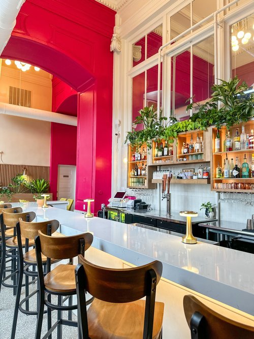 Modern bar with high ceilings, bright pink walls, a white countertop, wooden bar stools, potted plants, and a well-stocked back bar with glass shelving and mirrors. Warm lighting creates a welcoming atmosphere.
