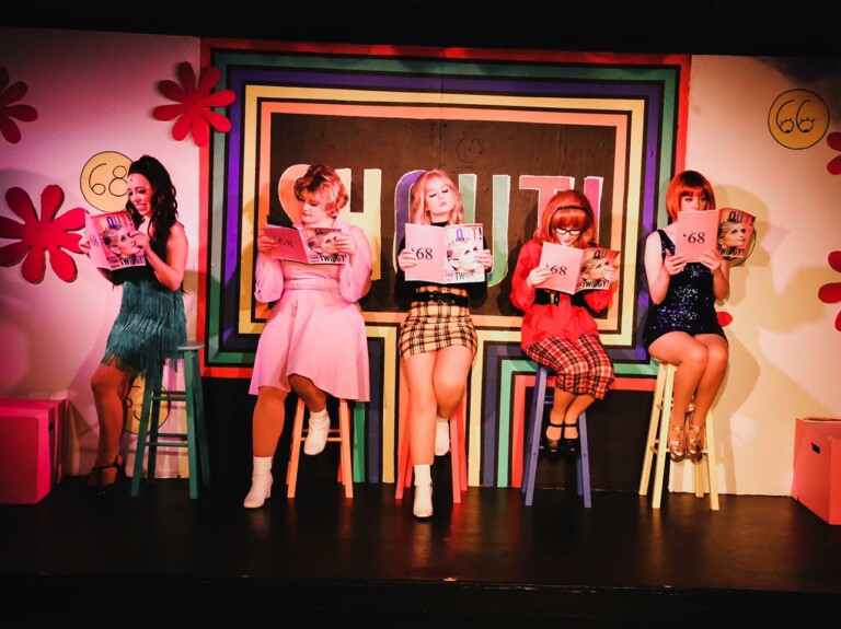 Five women dressed in colorful 1960s-inspired outfits sit on stools in front of a vibrant 