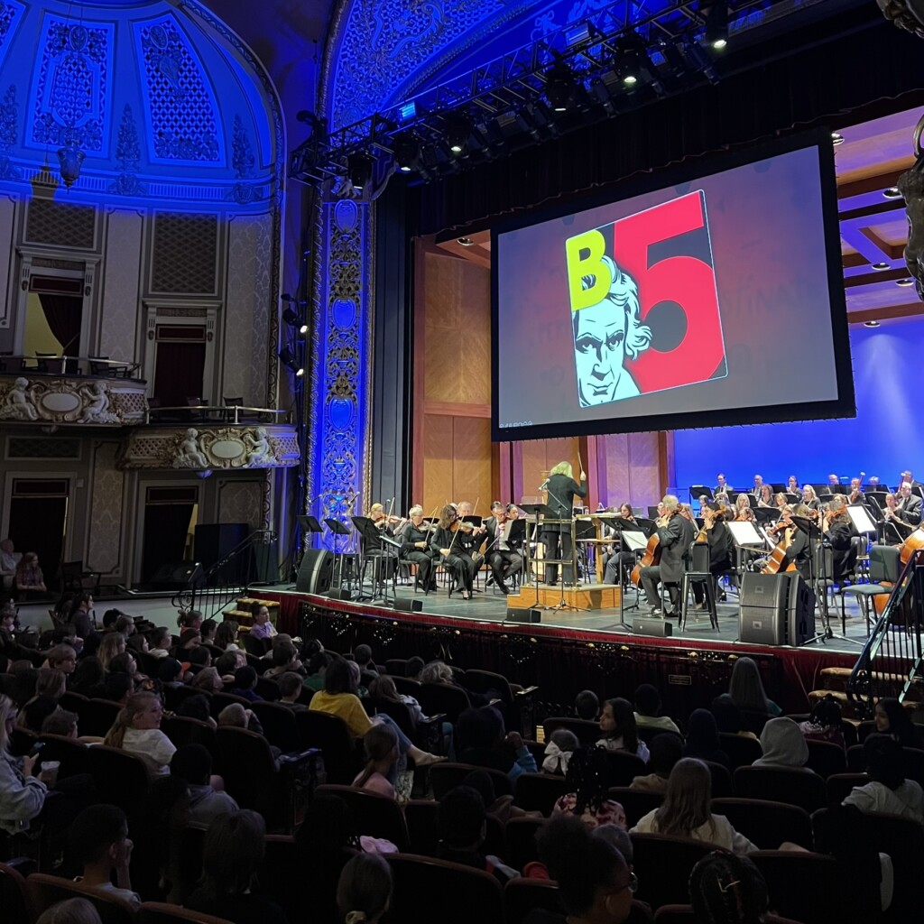 A symphony orchestra performs on stage in a grand theater, with a conductor leading and an audience watching. A screen above the stage displays an illustrated portrait and the text "B 5" in bold letters.