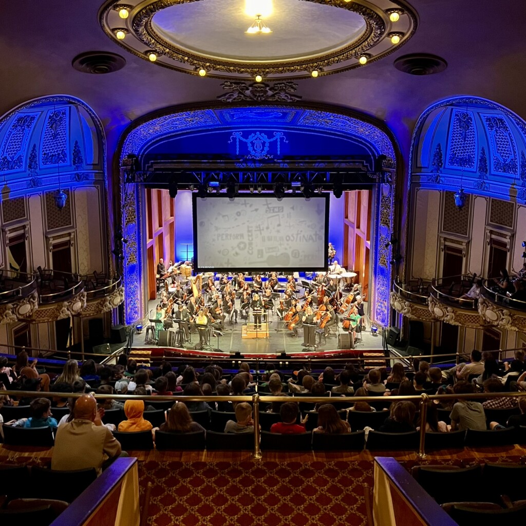 A symphony orchestra performs on a grand, ornate theater stage with blue-lit walls, as a large audience sits facing the musicians. The ceiling is domed and elaborately decorated.