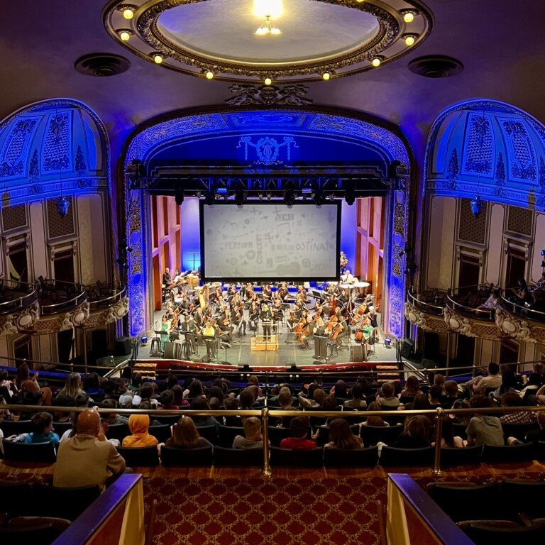 A symphony orchestra performs on a grand, ornate theater stage with blue-lit walls, as a large audience sits facing the musicians. The ceiling is domed and elaborately decorated.
