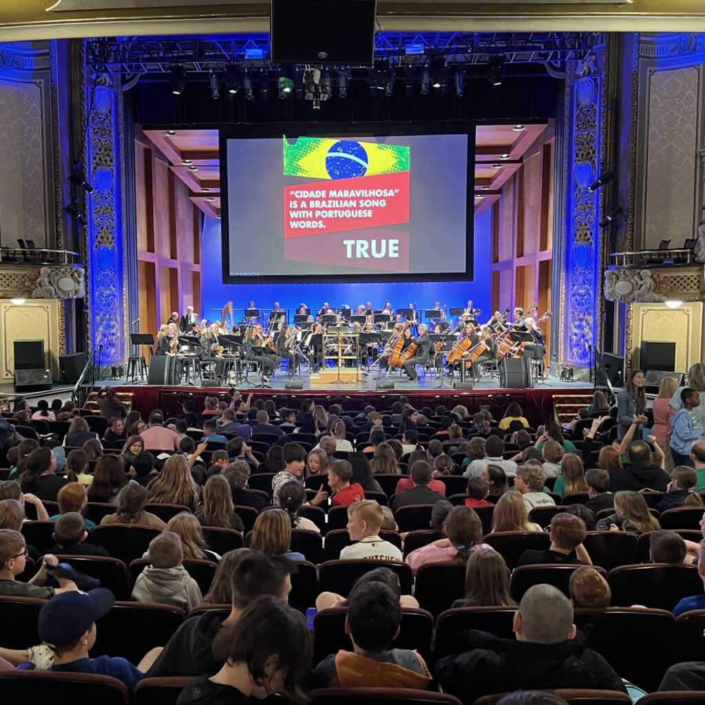 A full theater audience watches an orchestra perform on stage. A large screen above displays a Brazilian flag and a quiz statement about the song "Cidade Maravilhosa," with the answer "TRUE" highlighted.