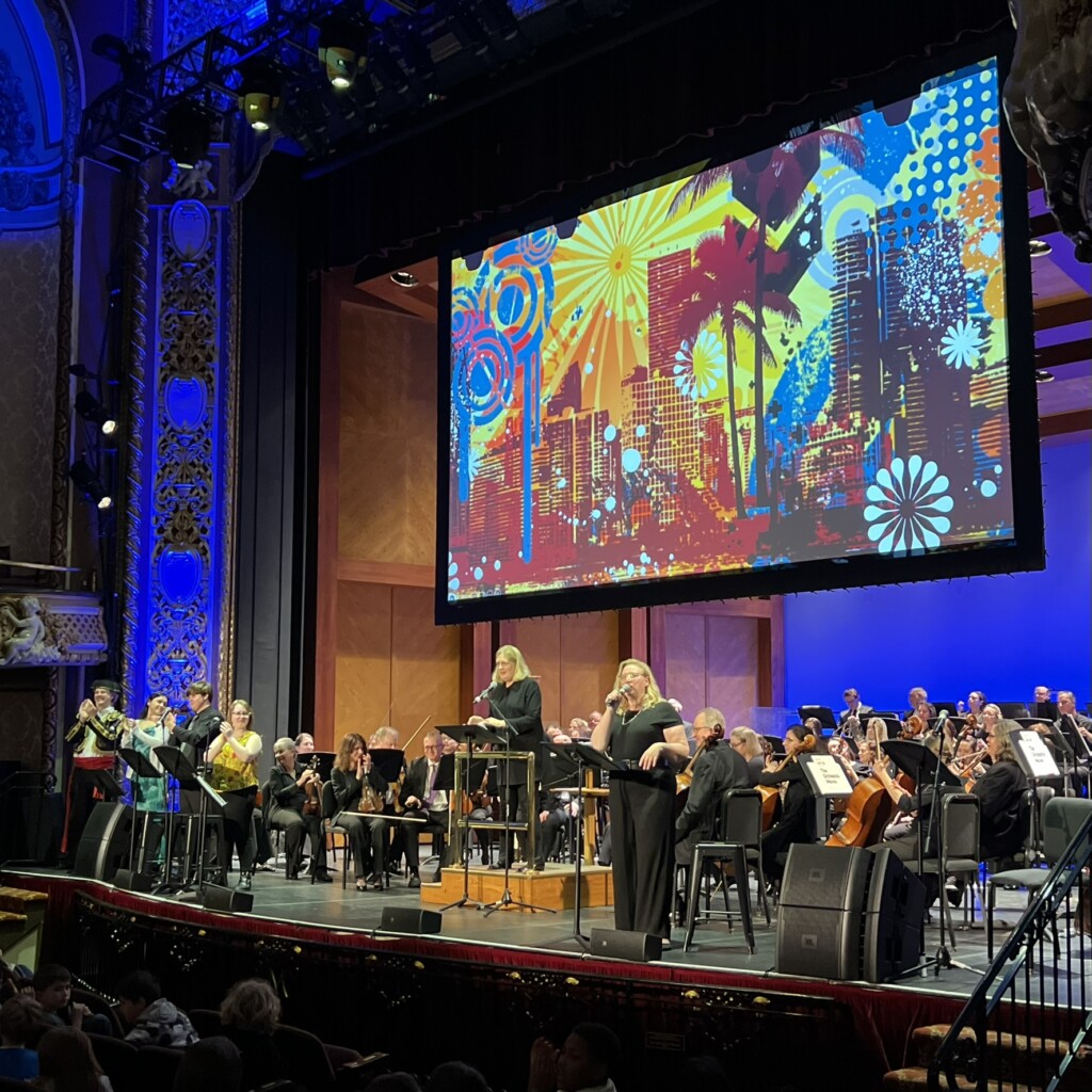 A symphony orchestra performs on a stage with a conductor and sign language interpreter. Behind them is a colorful, abstract cityscape projected on a large screen. The ornate theater is filled with an audience.