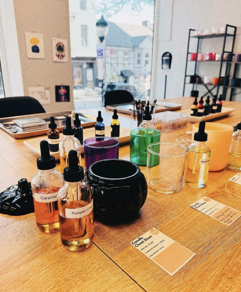 A tabletop is set with glass bottles of scented oils, empty candle jars, droppers, and colorful containers, ready for a candle-making workshop in a bright studio with large windows.