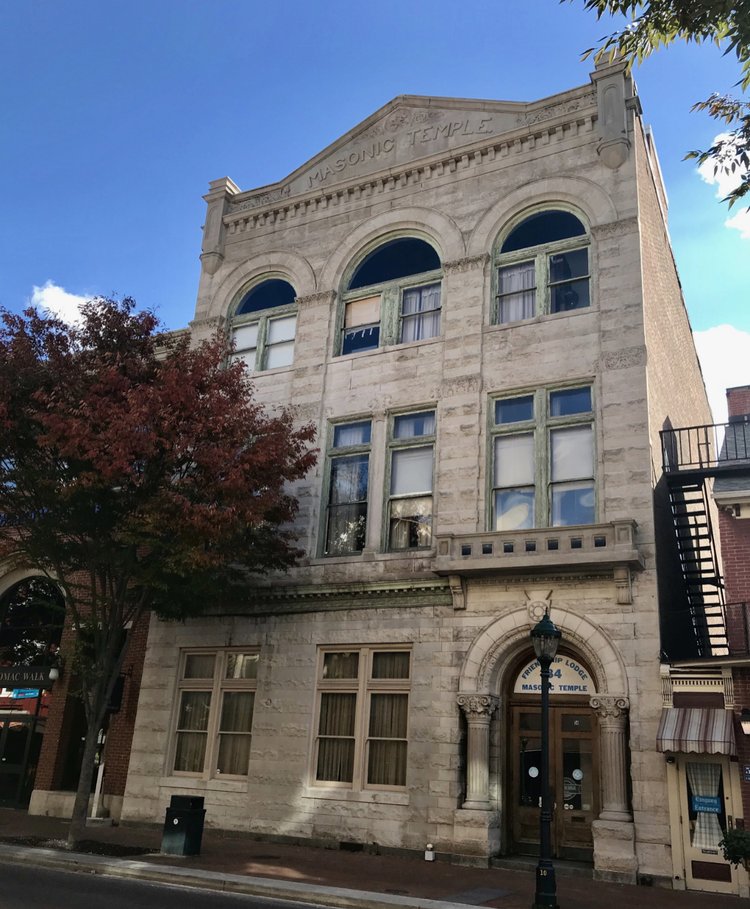 A three-story stone building with arched windows and “Masonic Temple” inscribed at the top. Trees flank the entrance, and a streetlamp stands on the sidewalk in front of the historic structure.
