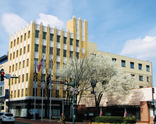 A four-story Art Deco building on a street corner, with blooming white trees in front and U.S. flags on poles. A pedestrian walks nearby under a blue sky with scattered clouds.