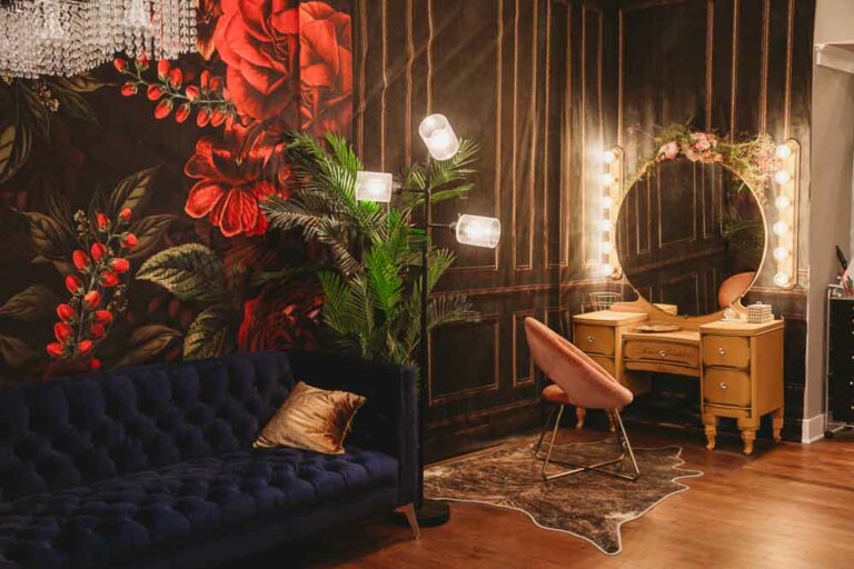 A stylish room with a dark floral mural, blue velvet sofa, tall floor lamp, a yellow vanity with round mirror framed by lights, a pink chair, a faux fur rug, and a leafy plant on wood flooring.