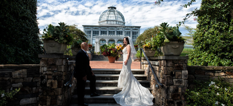 A bride in a white gown and a groom in a black suit pose on stone steps outside a glass conservatory, surrounded by greenery and potted plants under a partly cloudy sky.