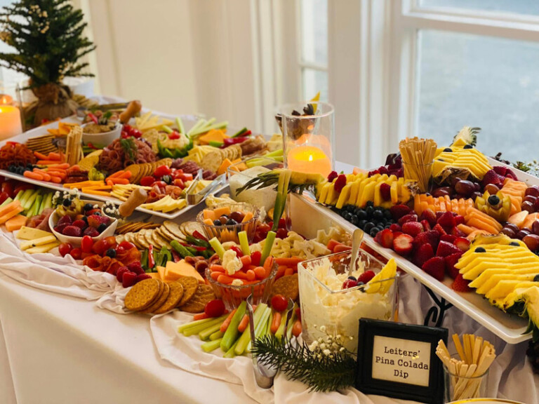 A colorful grazing table with assorted fruits, vegetables, crackers, cheeses, dips, and breadsticks, decorated with candles and greenery, set near large windows with natural light. A sign reads 