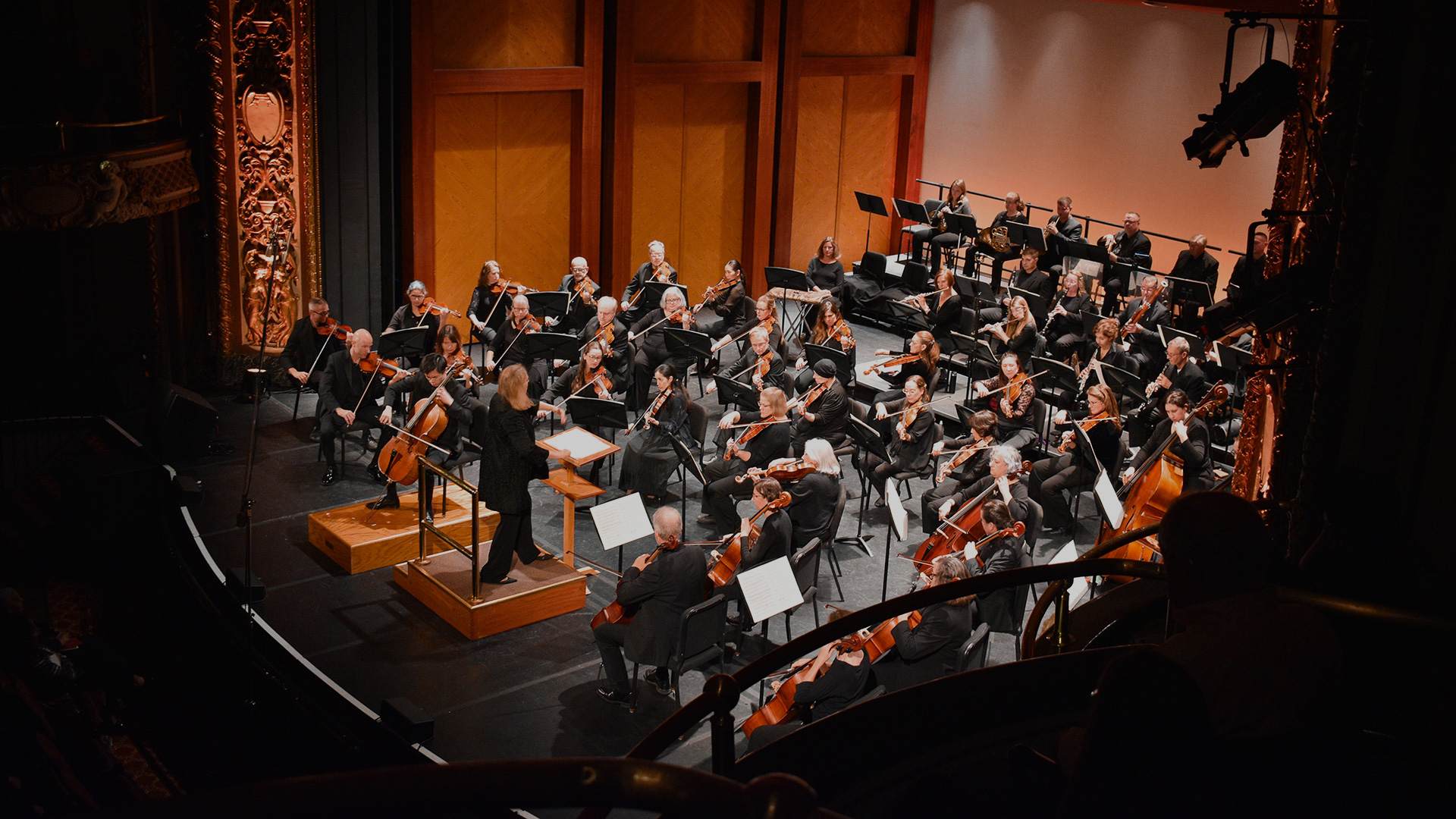 A conductor leads an orchestra of musicians playing various string, brass, and woodwind instruments on a stage in a grand concert hall, viewed from a high balcony.