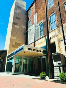 Entrance of a modern building called "The Updegraff," featuring a glass doorway, beige panels, old brick walls, large windows, and a tall metal sculpture beside a paved walkway under a bright blue sky.