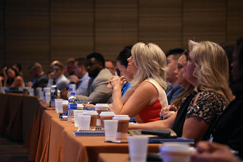 A group of people sits at long tables, facing forward and listening attentively during a conference or seminar. Notebooks, coffee cups, and water bottles are on the tables. The room appears dimly lit.