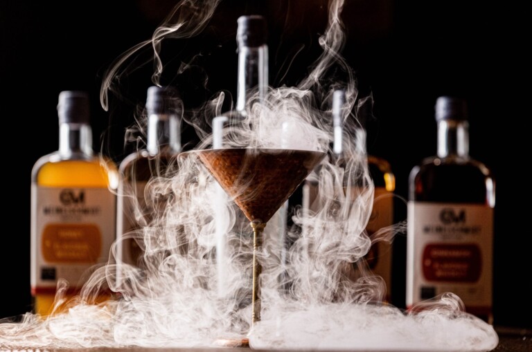 A smoky cocktail in a martini glass is surrounded by swirling vapor, with several bottles of amber-colored liquor blurred in the background against a dark backdrop.