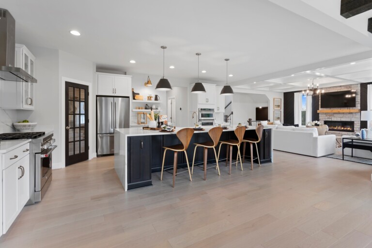 Modern open-concept kitchen with a large island, four wooden bar stools, pendant lights, stainless steel appliances, white cabinets, and a connected living room with a fireplace and TV.