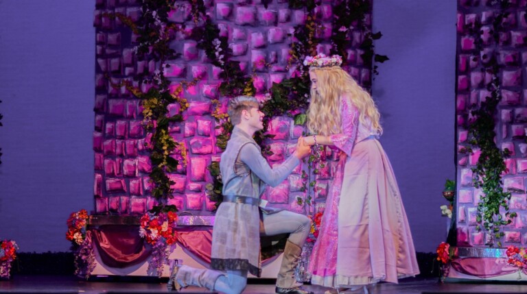 A young man kneels and holds hands with a woman in a long pink dress and flower crown, performing on stage with a floral, ivy-covered backdrop and purple lighting.