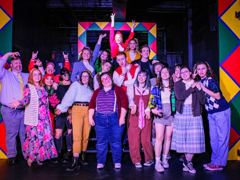 A large group of people of various ages pose in colorful, lively outfits on a stage with a vibrant geometric background, some making playful gestures and smiling enthusiastically at the camera.