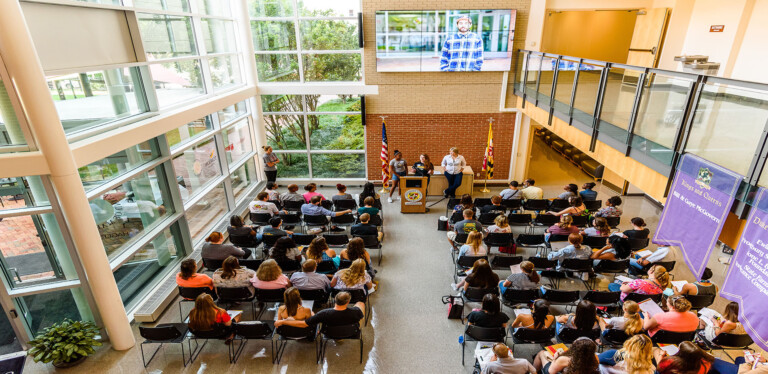 A group of people sit in rows facing a podium in a bright, modern atrium with large windows. Three speakers stand at the podium, and a large screen above displays a person's portrait. Flags and a purple banner are visible.