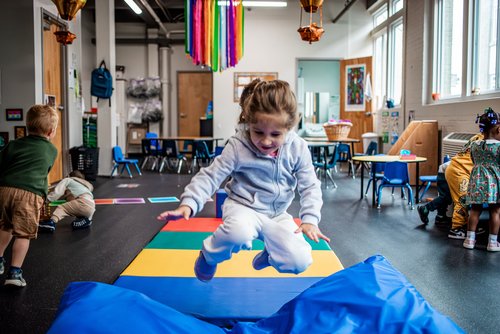 A young girl in a gray tracksuit jumps energetically onto blue soft play mats in a colorful classroom, while other children play in the background.