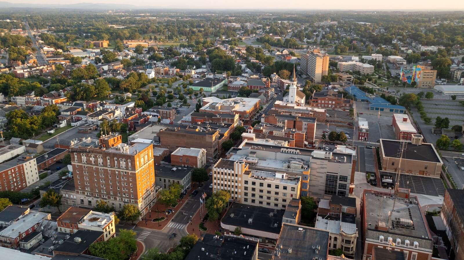 Aerial view of a small city with a mix of historic and modern buildings, green spaces, and tree-lined streets under a clear sky during sunset.