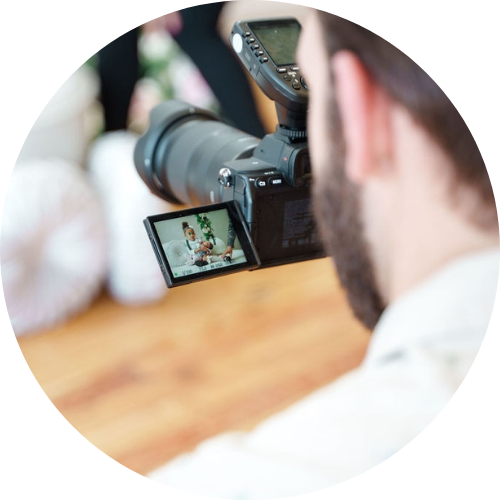 A person is photographing or filming with a digital camera, capturing an image of another person sitting on a couch, as seen on the camera’s flip-out screen. The scene is indoors with a blurred background.
