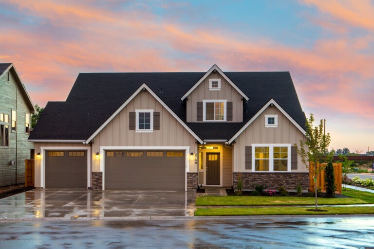 A two-story suburban house with beige siding, stone accents, and three-car garage, photographed at sunset with a wet driveway and manicured lawn.