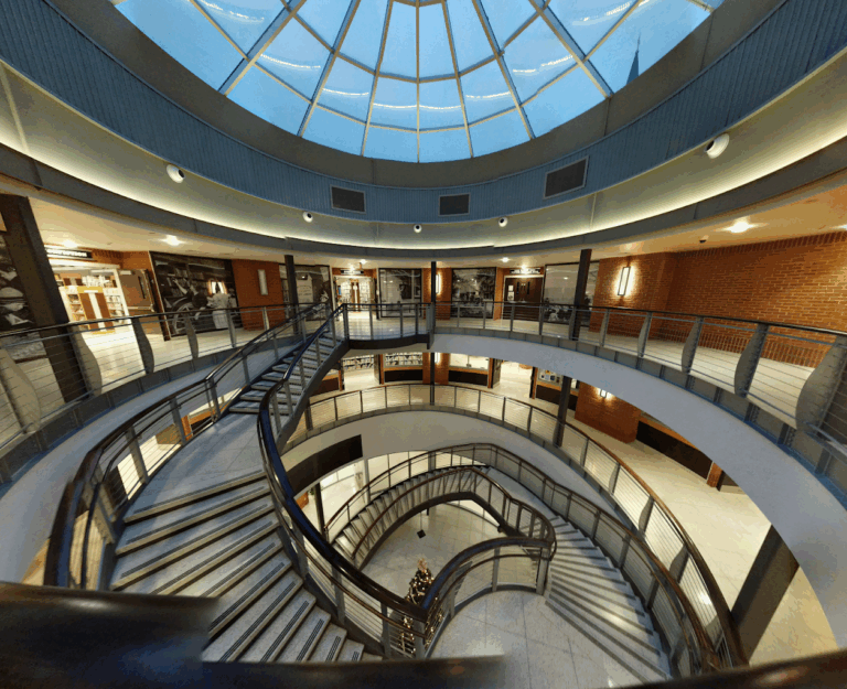 A wide-angle view of a multi-level atrium with a spiral staircase, glass dome ceiling, curved railings, and a decorated Christmas tree on the ground floor. Warm lighting highlights the modern architecture.