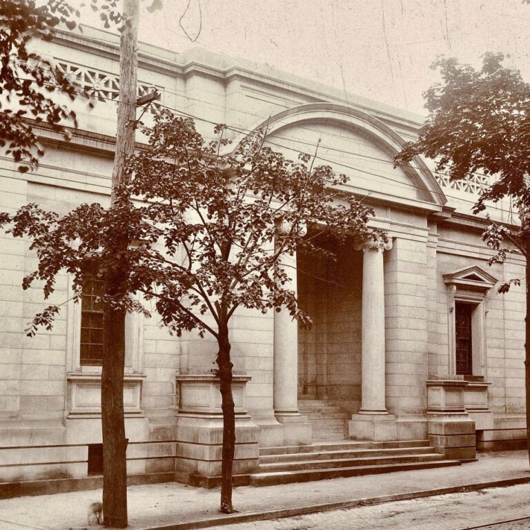 A sepia-toned photo of a neoclassical building with columns, arched windows, and stone steps, partially obscured by trees along a sidewalk.